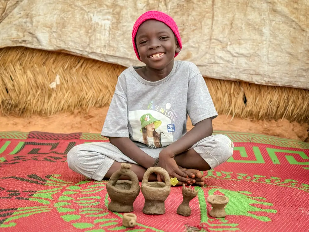 Rayan, de 7 años, es una de las miles de niños y niñas sudaneses que viven en el campo de tránsito de Adré. Posa orgullosa frente a las tazas y teteras que fabrica con barro que recoge después de la lluvia.