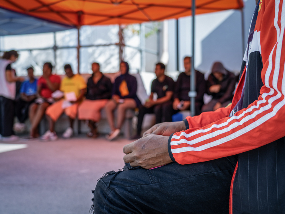 En la foto, un médico de MSF con ropa protectora, se inclina para estar al nivel de la cama de su paciente, una pequeña con sarampión recostada en la coma, cubierta por una manta y con una sonda nasal. Al lado de la pequeña, está su madre.