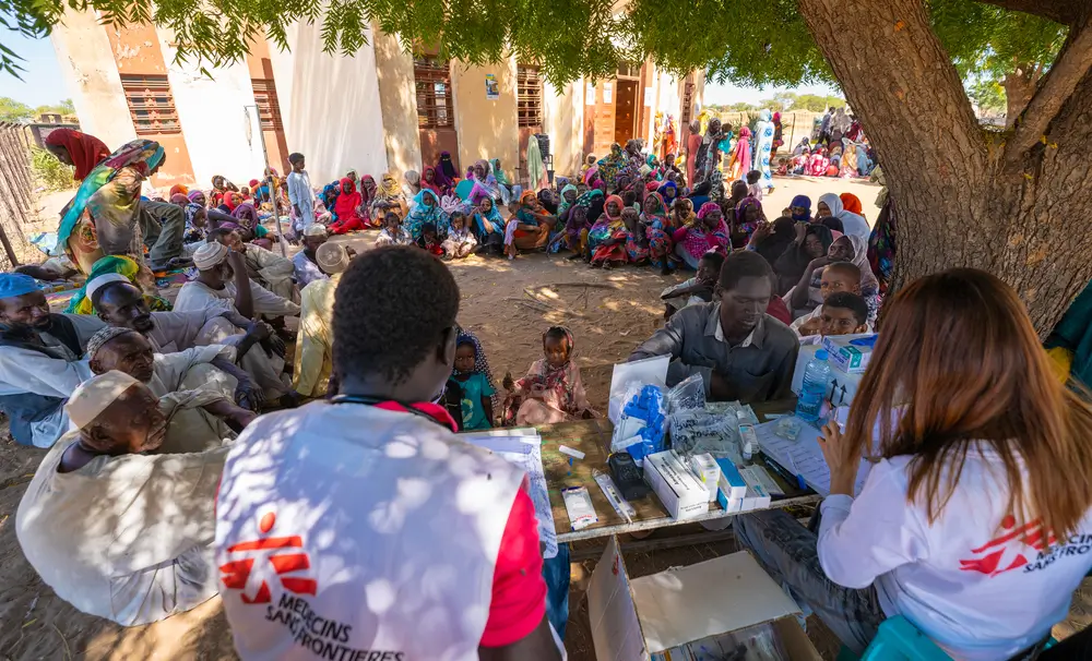Una multitud de retornados y refugiados espera en fila bajo un árbol para recibir atención del equipo de la clínica móvil de MSF en el asentamiento informal de Jerbana.